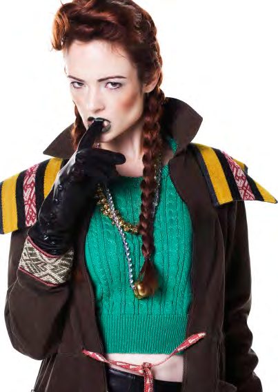 Model with a long braid in a green crop sweater and brown anorak with Peruvian trim, a gloved finger pressed to her lips
