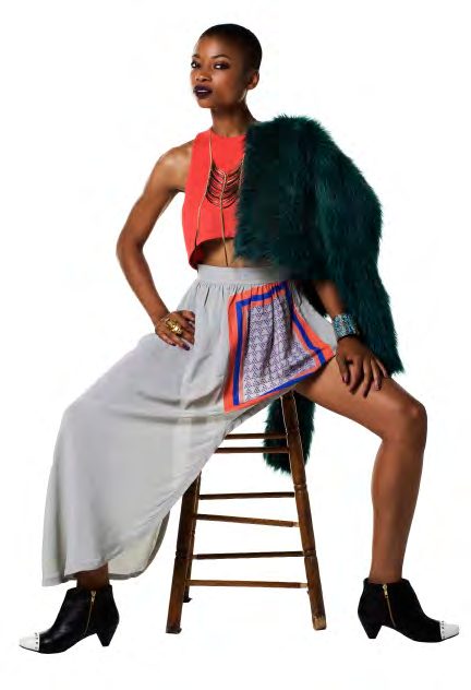 Model seated on a wooden stool in a coral crop top, long grey skirt and a dark green faux fur jacket