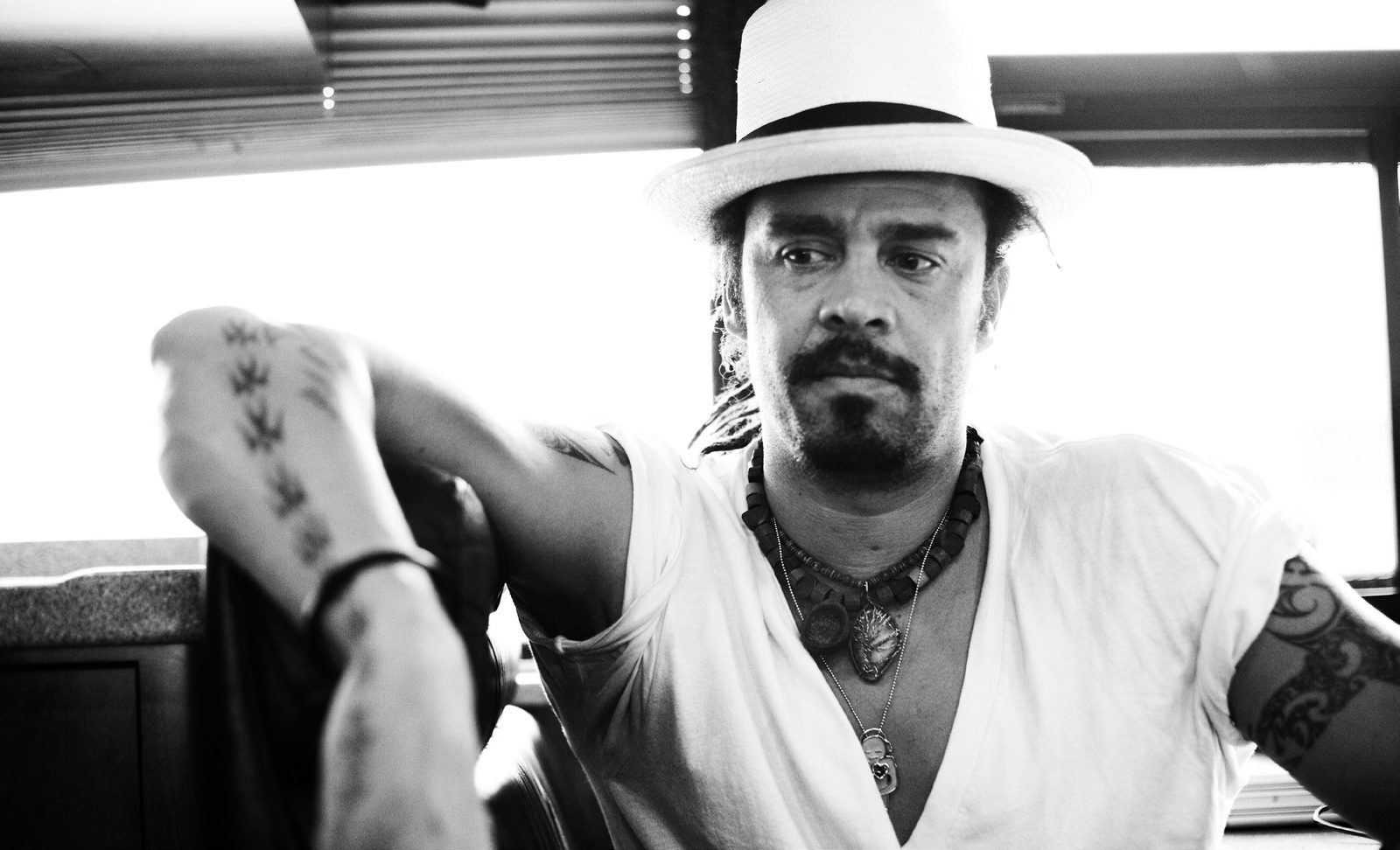Black and white photo of Michael Franti in a white fedora and V-neck tee leaning on a window ledge, tattoos on his forearms