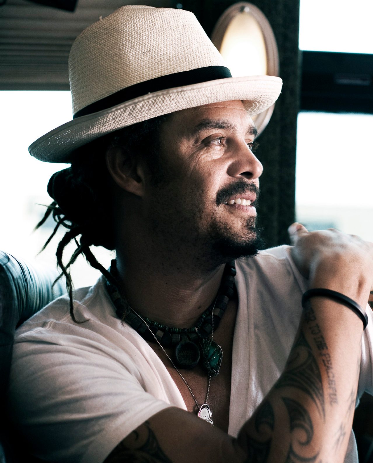 Michael Franti smiling in a straw fedora with dreadlocks, beaded necklaces and a tattooed arm resting on a window ledge