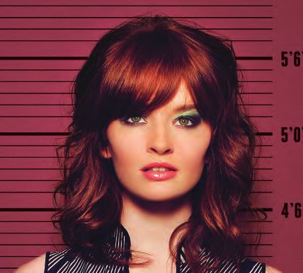 Mug shot of a model with long auburn hair, heavy bangs and green eyeshadow against a crimson height chart