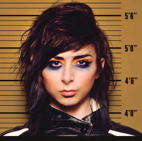 Mug shot of a model with tousled dark hair and a smudged blue and orange smoky eye against a mustard height chart