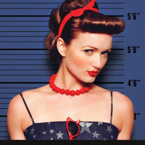 Mug shot of a pin-up model with victory rolls, a red bandana, red lips and a red bead necklace against a blue height chart