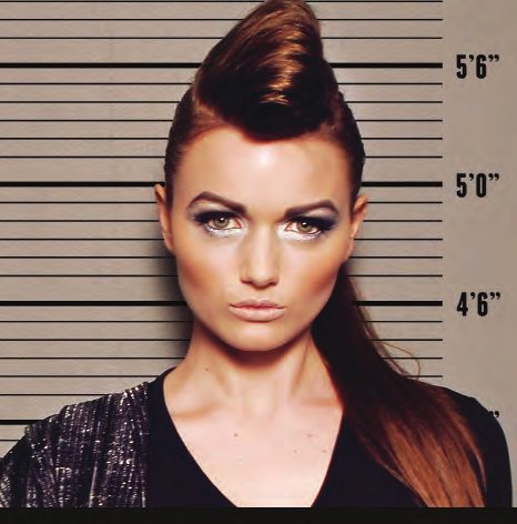 Mug shot of a model with a sculpted auburn pompadour and metallic eye makeup against a taupe height chart