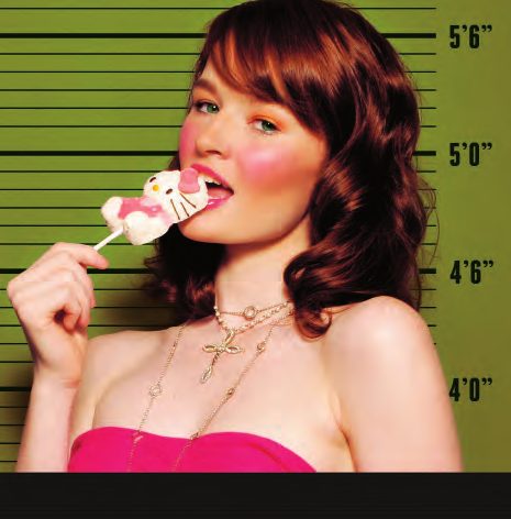 Mug shot of a model with auburn curls and bright pink blush licking a Hello Kitty lollipop against a lime height chart