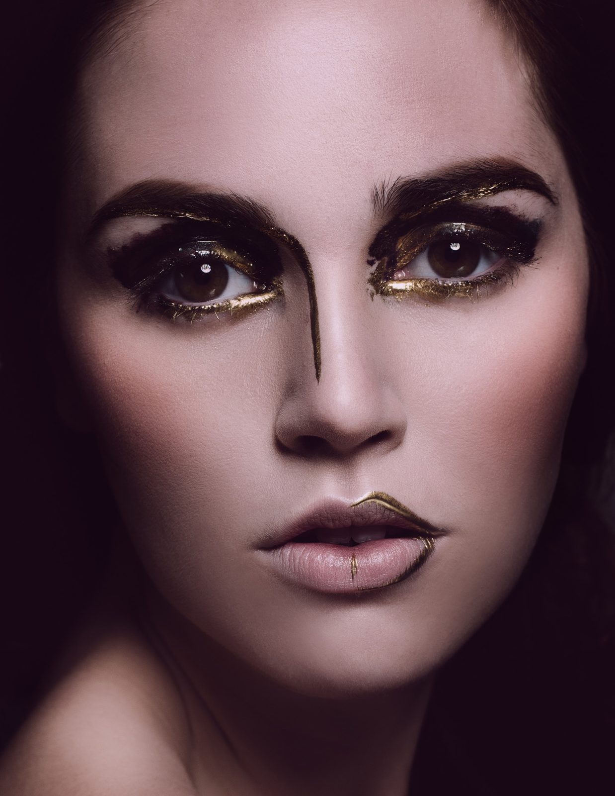 Model with dark smoky eyes, gold liner, a gold stripe down the bridge of her nose and a sculpted bronze lip