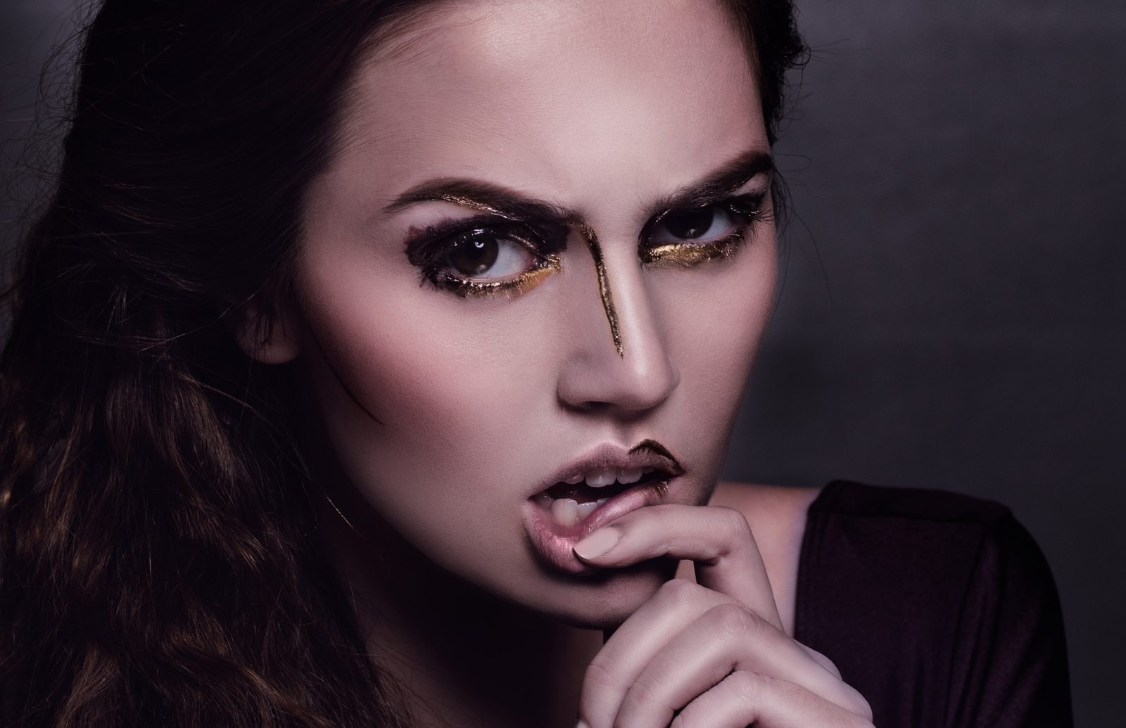 Model with heavy black and gold eye makeup, a gold line down her nose and a bronze lip, finger pressed to her mouth
