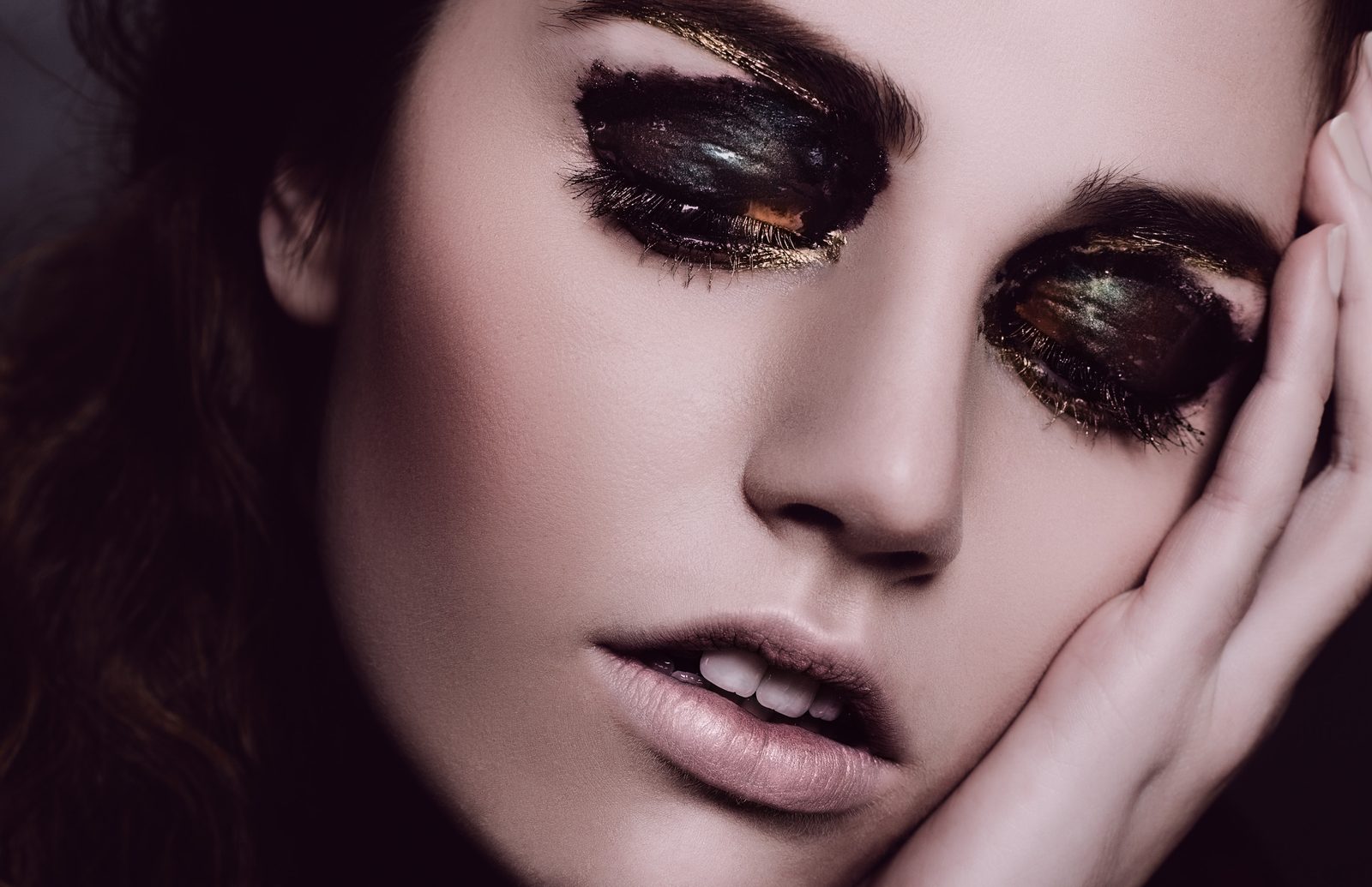 Close-up of a model with eyes closed under glossy black lacquered lids edged in gold, hand resting against her cheek
