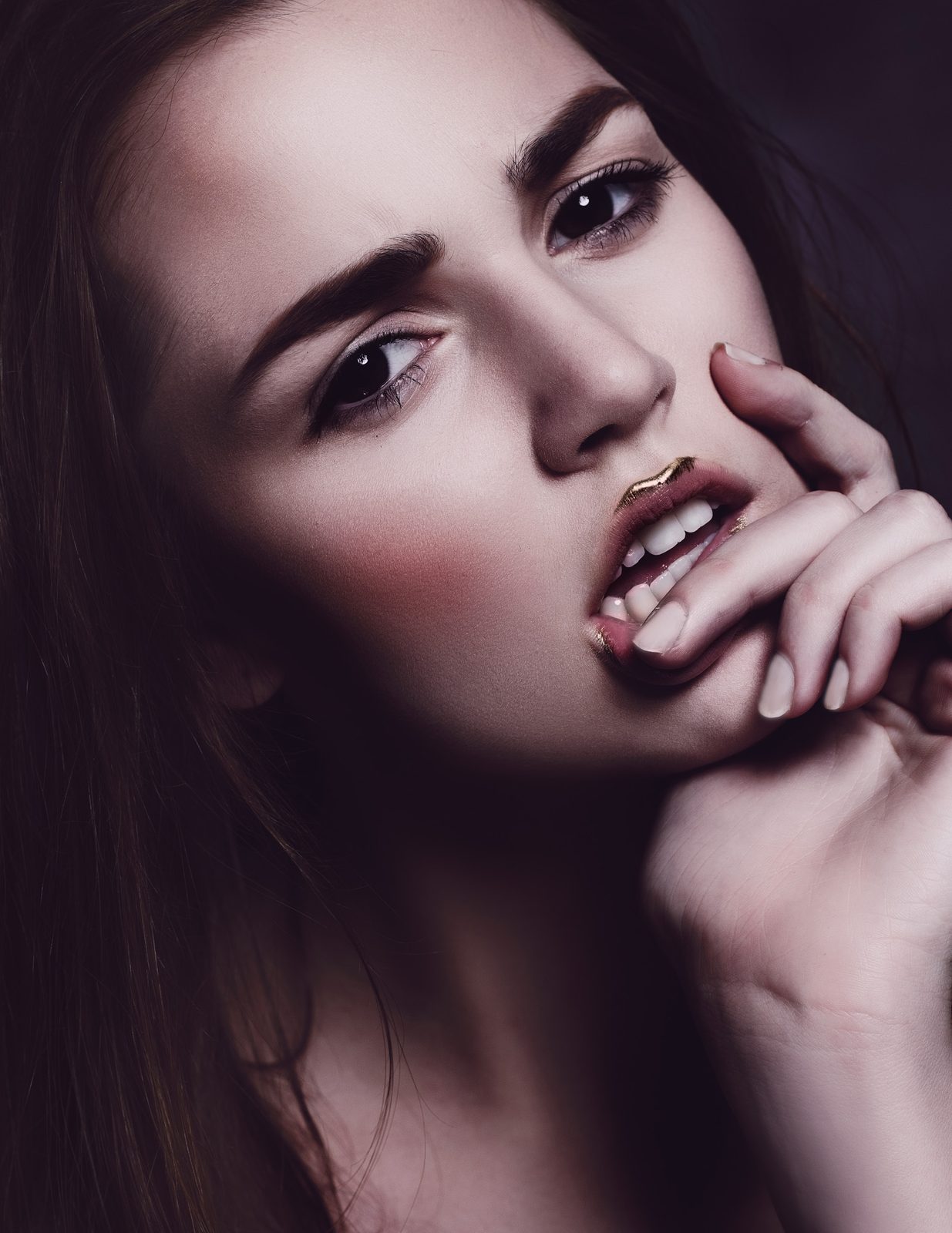 Model with long dark waves and dark lips biting her fingertip, gold flecks on her lashes
