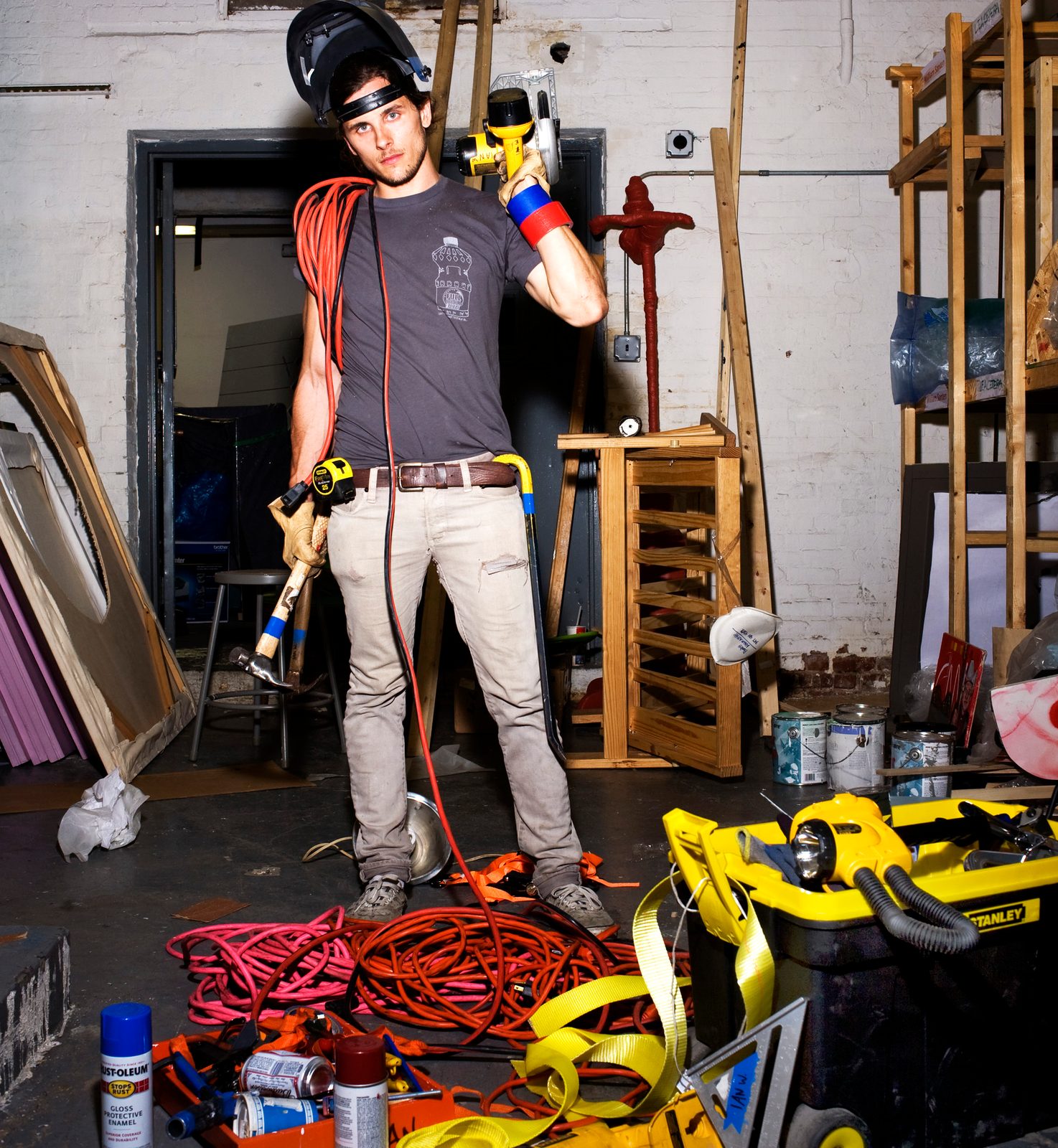 Ian Warren in a welding helmet and tool belt holding a power tool in a cluttered workshop