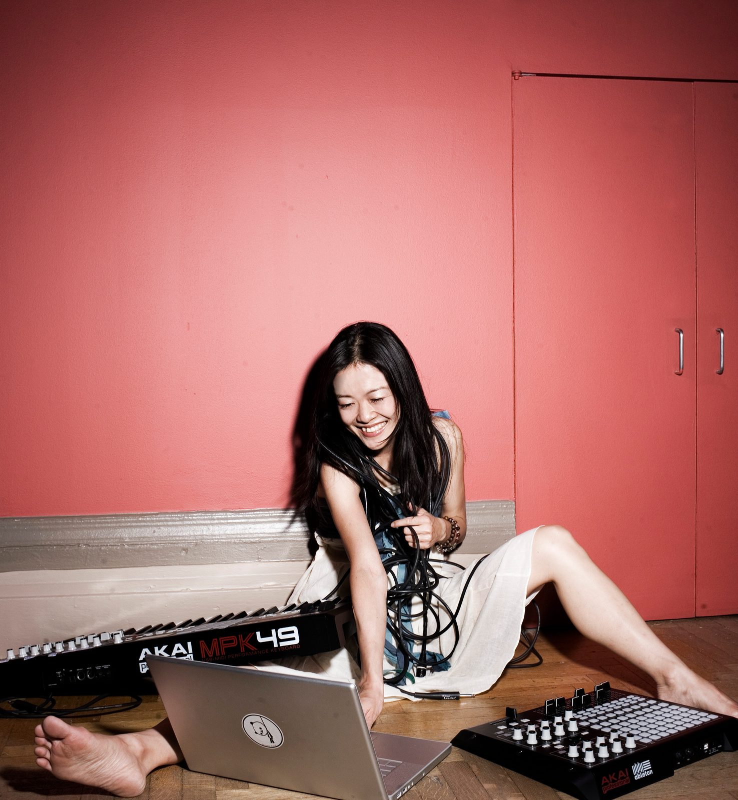 Yoko K. sitting barefoot on the floor against a pink wall surrounded by a laptop, MIDI keyboard and tangled cables