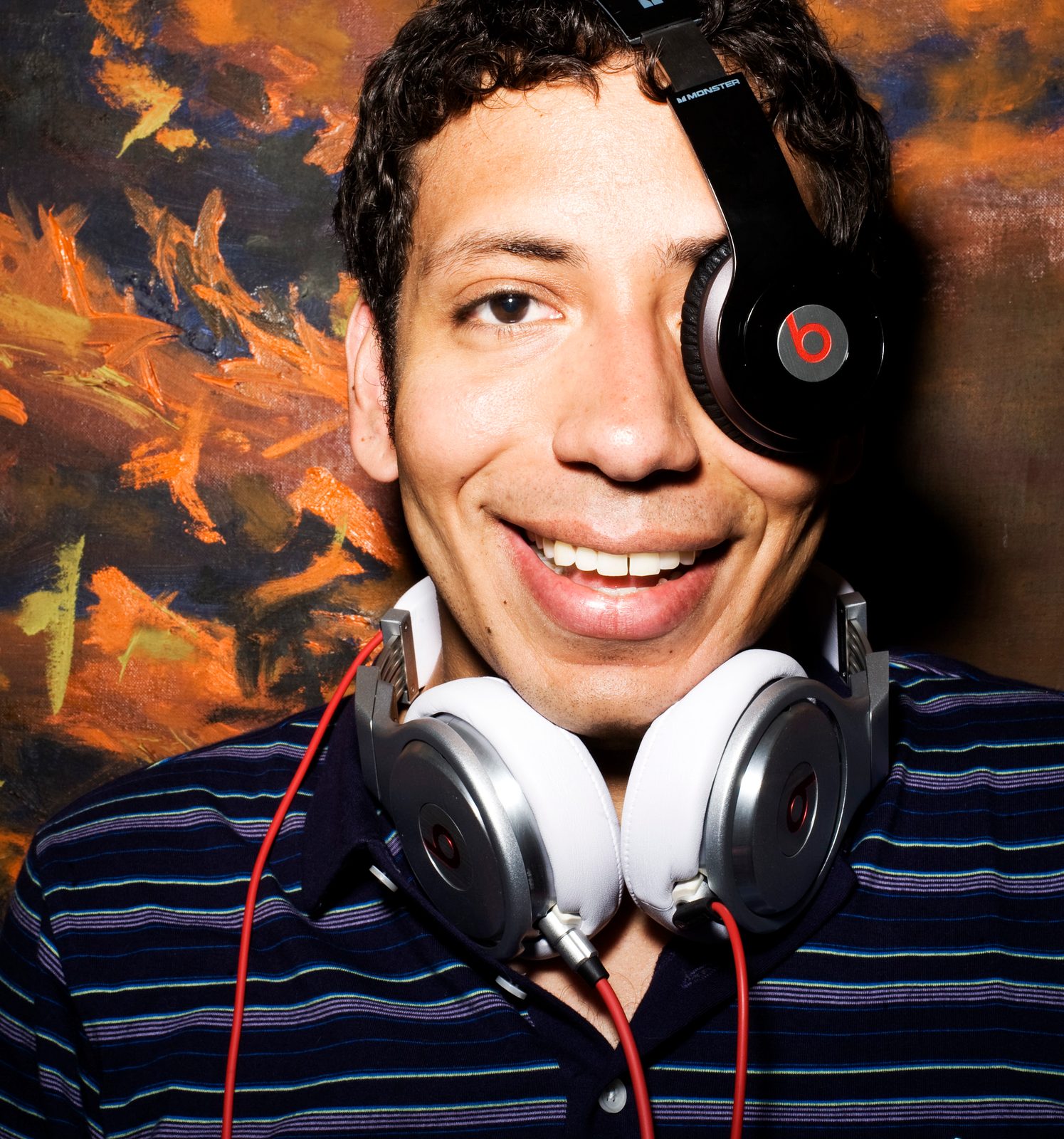 Derek James grinning with one headphone over his eye and another pair around his neck in front of a fiery painting