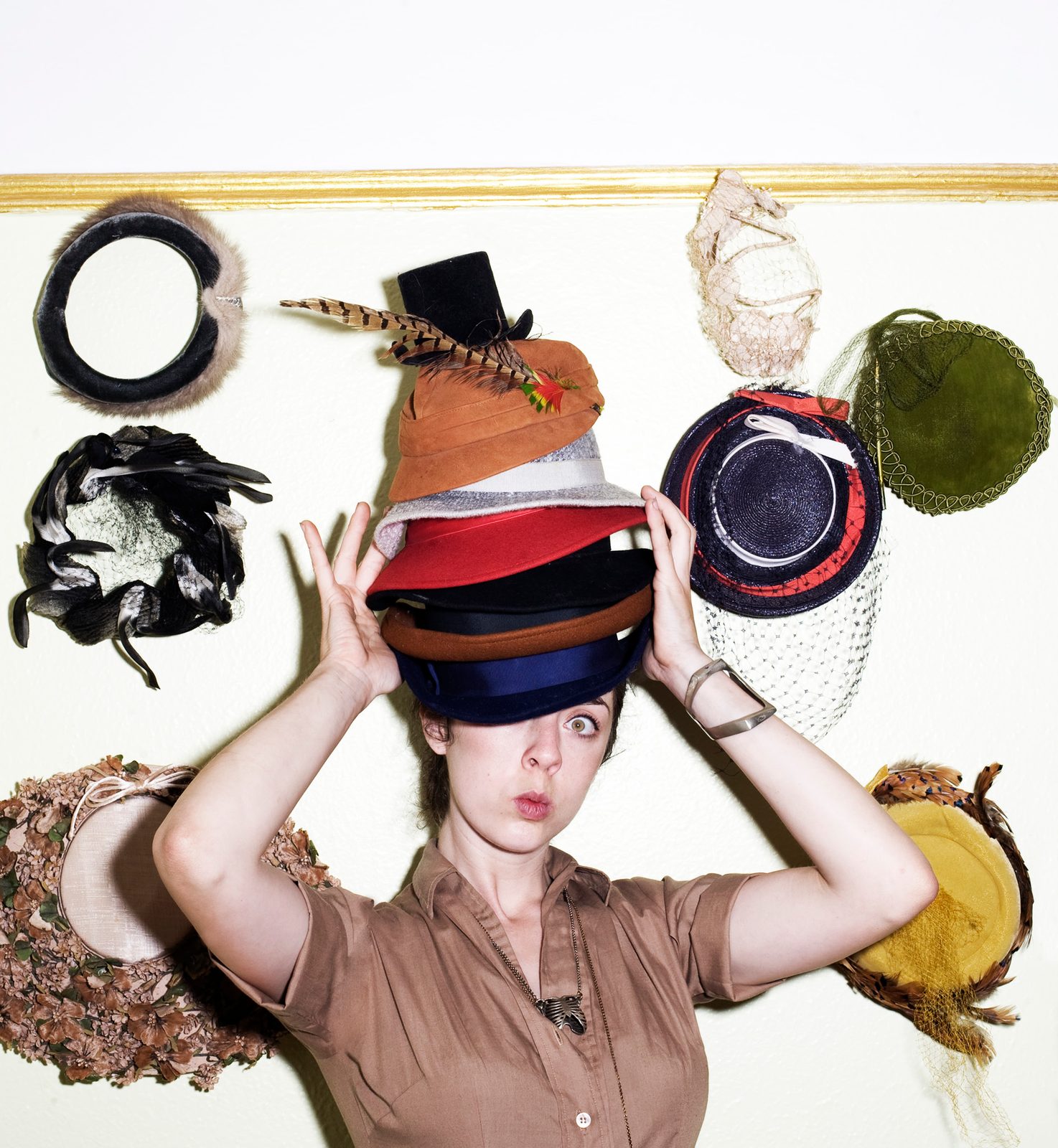 Lillian Crowe balancing a towering stack of vintage hats on her head against a wall hung with more hats and hair pieces