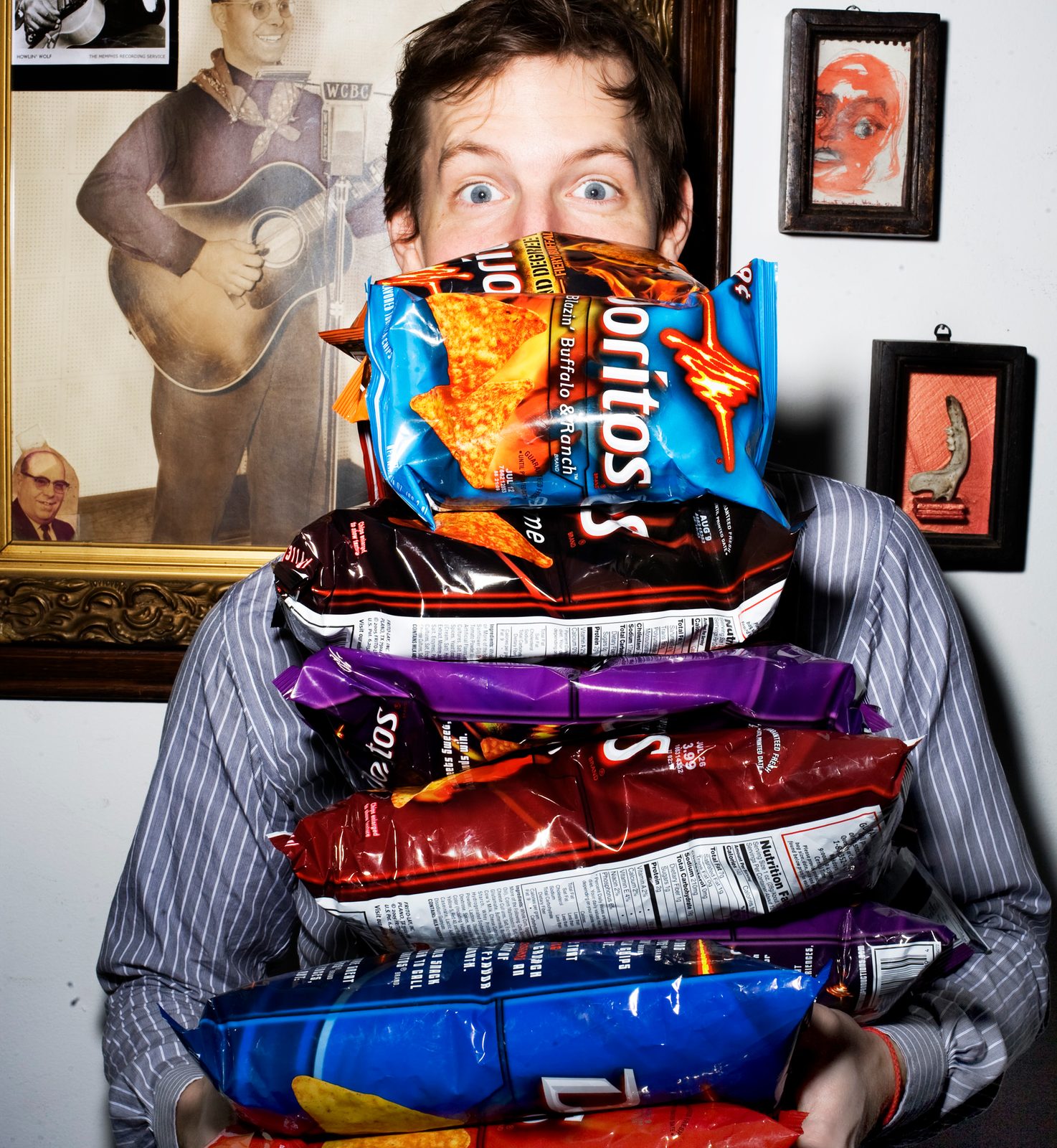 Chris Eldridge peering over an armful of stacked Doritos bags in a room hung with framed pictures