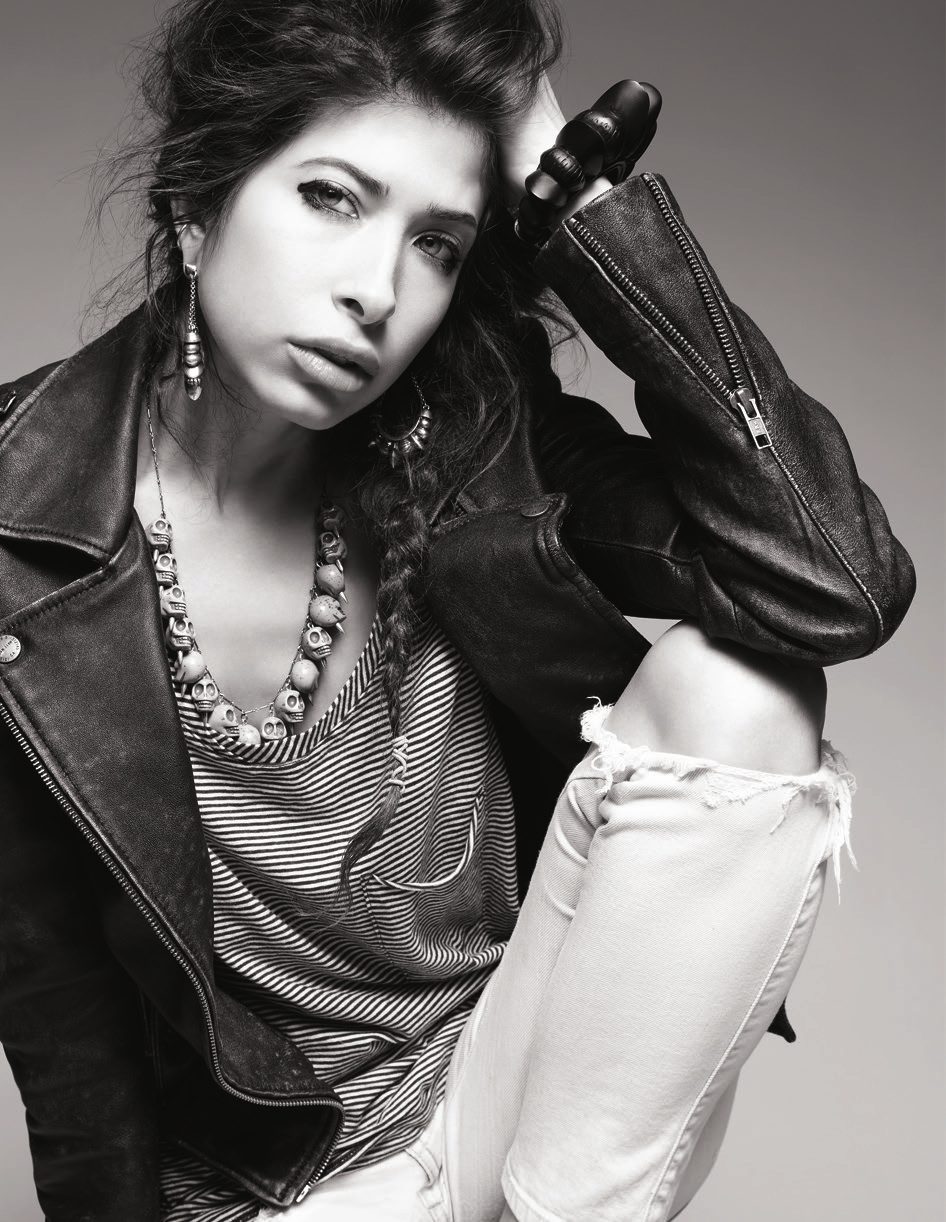 Black and white portrait of Pamela Love in a leather jacket, striped top and layered necklaces, one knee raised
