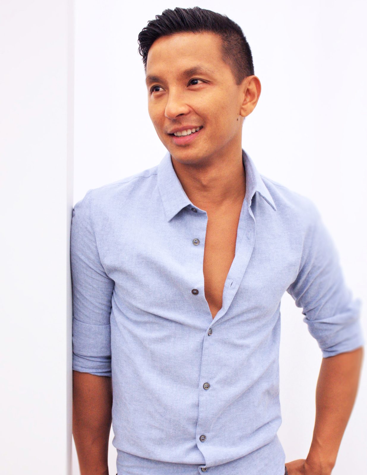 Prabal Gurung smiling in an open-collar light blue shirt with one hand on his hip against a white wall