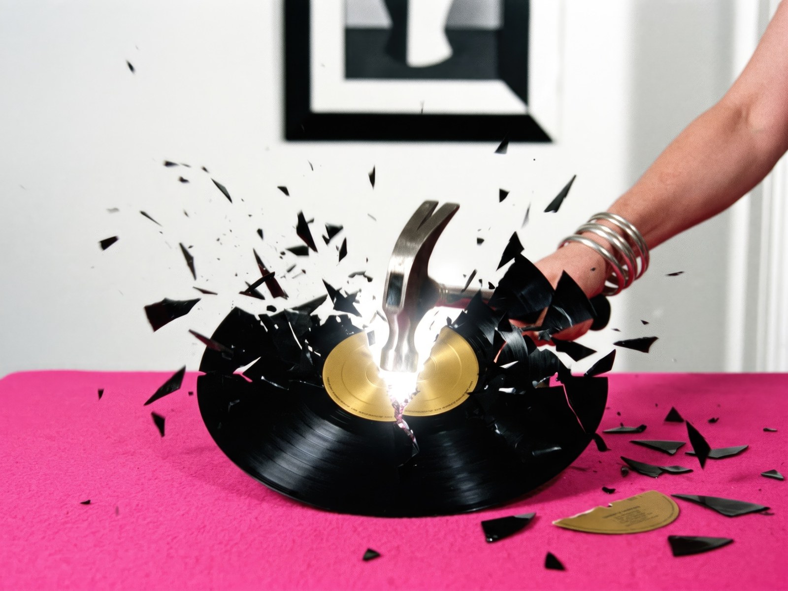 A hand with silver bangles smashes a black vinyl record with a hammer, shards flying across a hot pink table