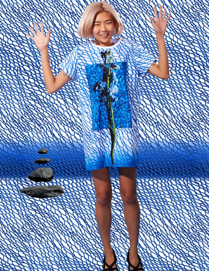 Model with a platinum bob in a blue scribble print shift dress with a photo print panel, hands raised, posed against a matching blue scribble backdrop with stacked stones