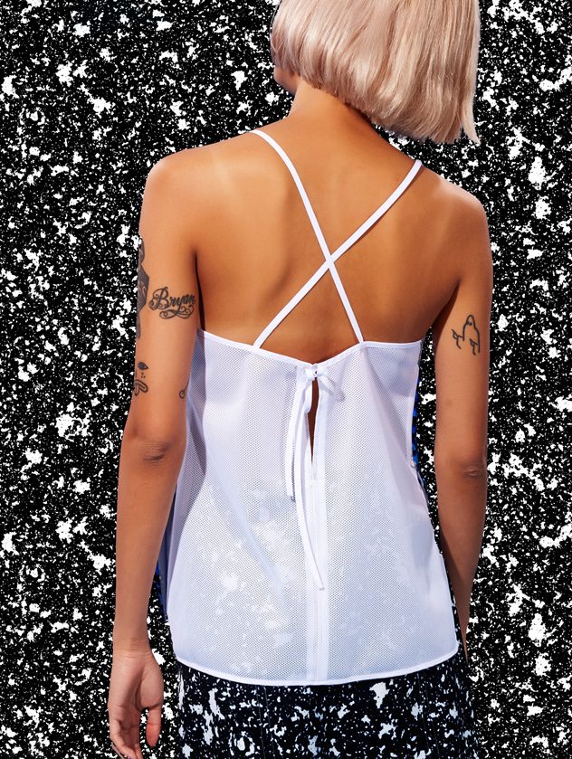 Back view of a model with a platinum bob in a white cami top with crossed straps against a black and white speckled backdrop