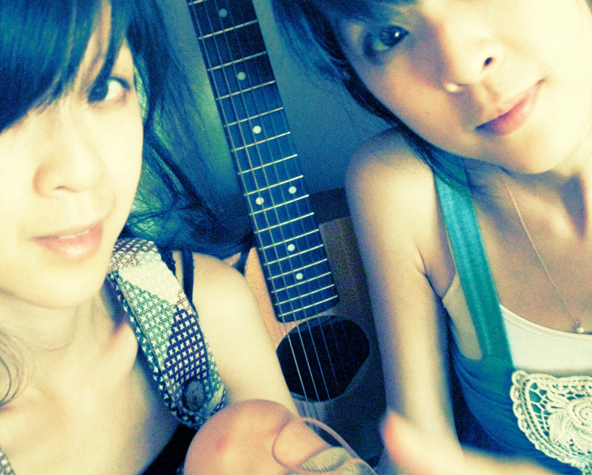 Selfie of twin sisters Emily and Susan Hsu with dark bangs leaning together over an acoustic guitar