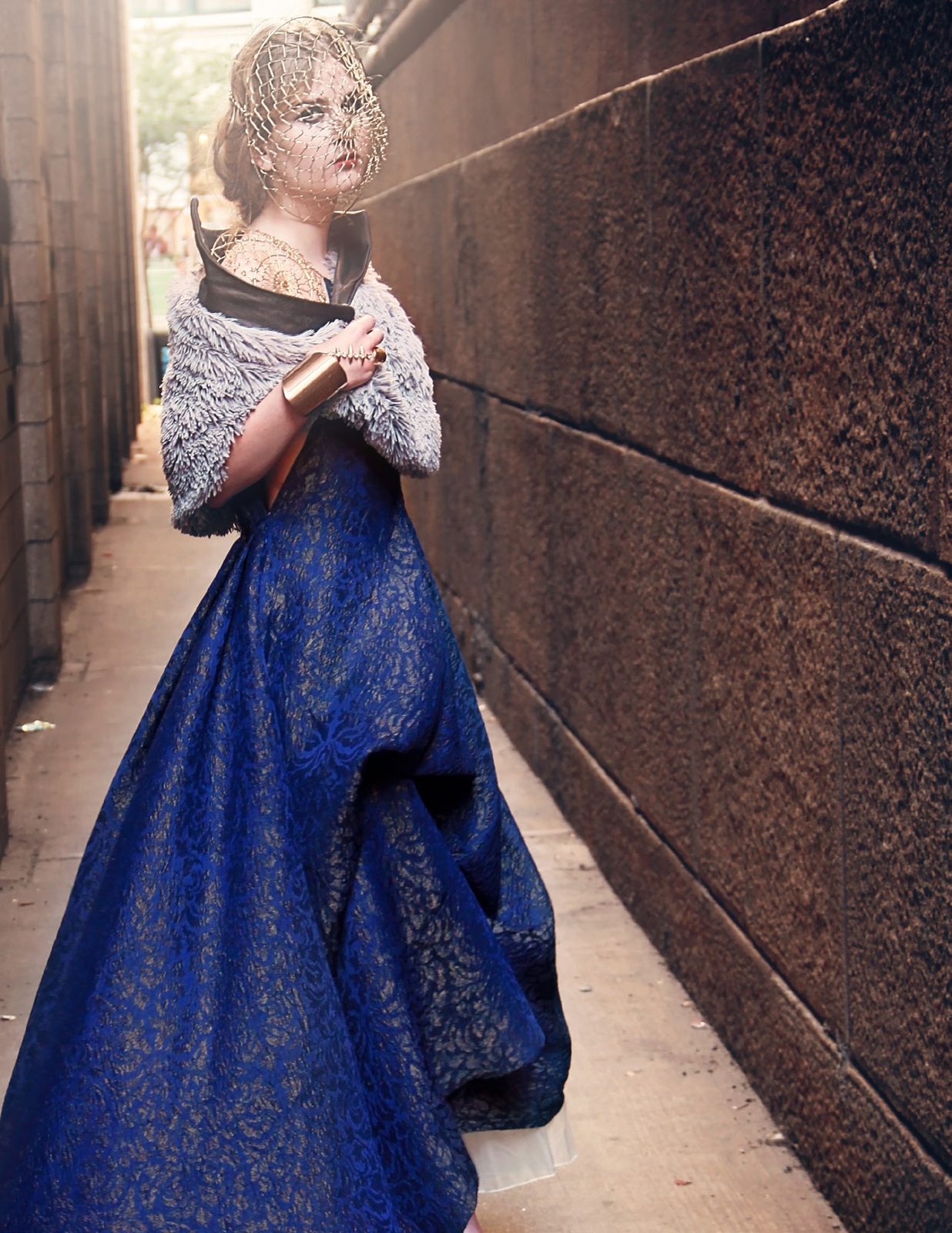 Model in a midnight blue brocade gown with a fur-trimmed capelet and gold headpiece stands in a narrow brick alley
