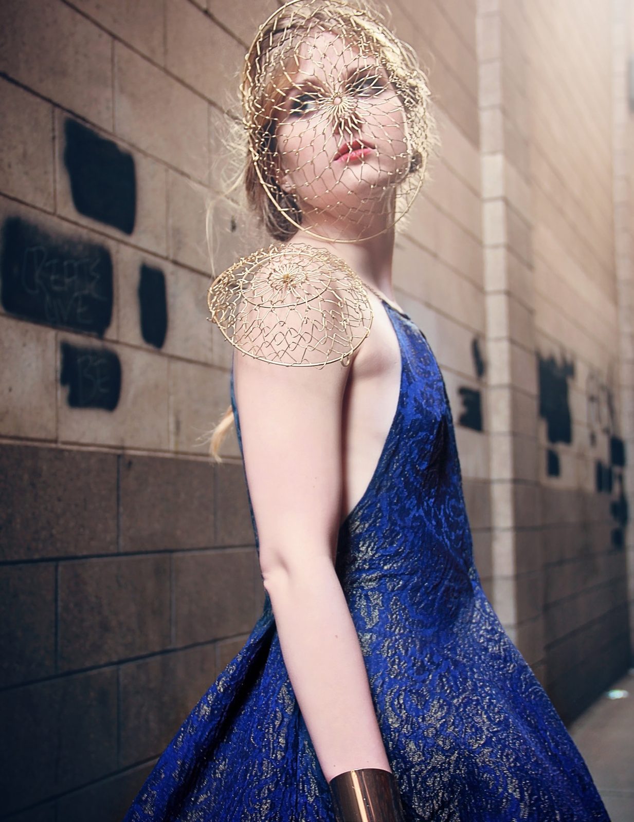 Model in a midnight blue brocade gown with a gold wire shoulder piece and caged headpiece leans against a brick wall
