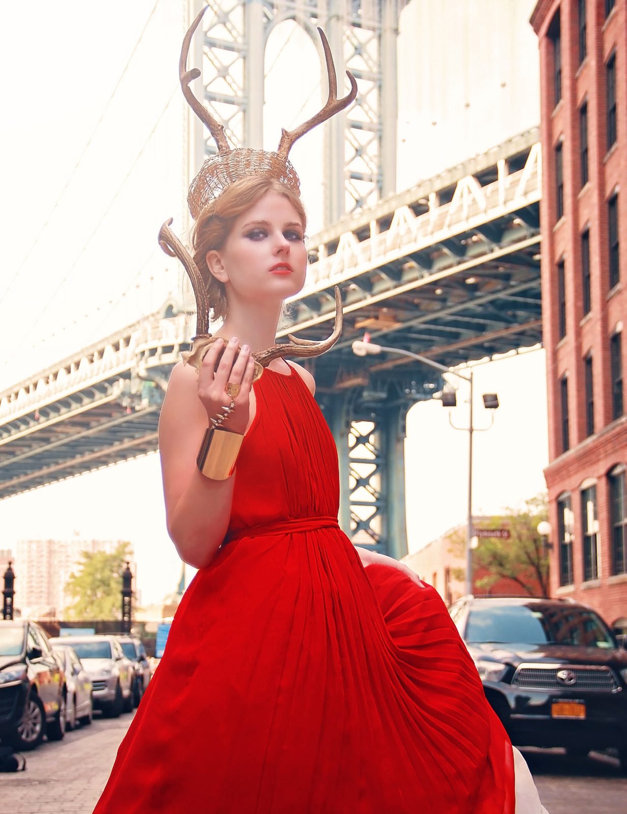 Model in a flowing red gown and antler headdress stands on a city street under a large steel bridge