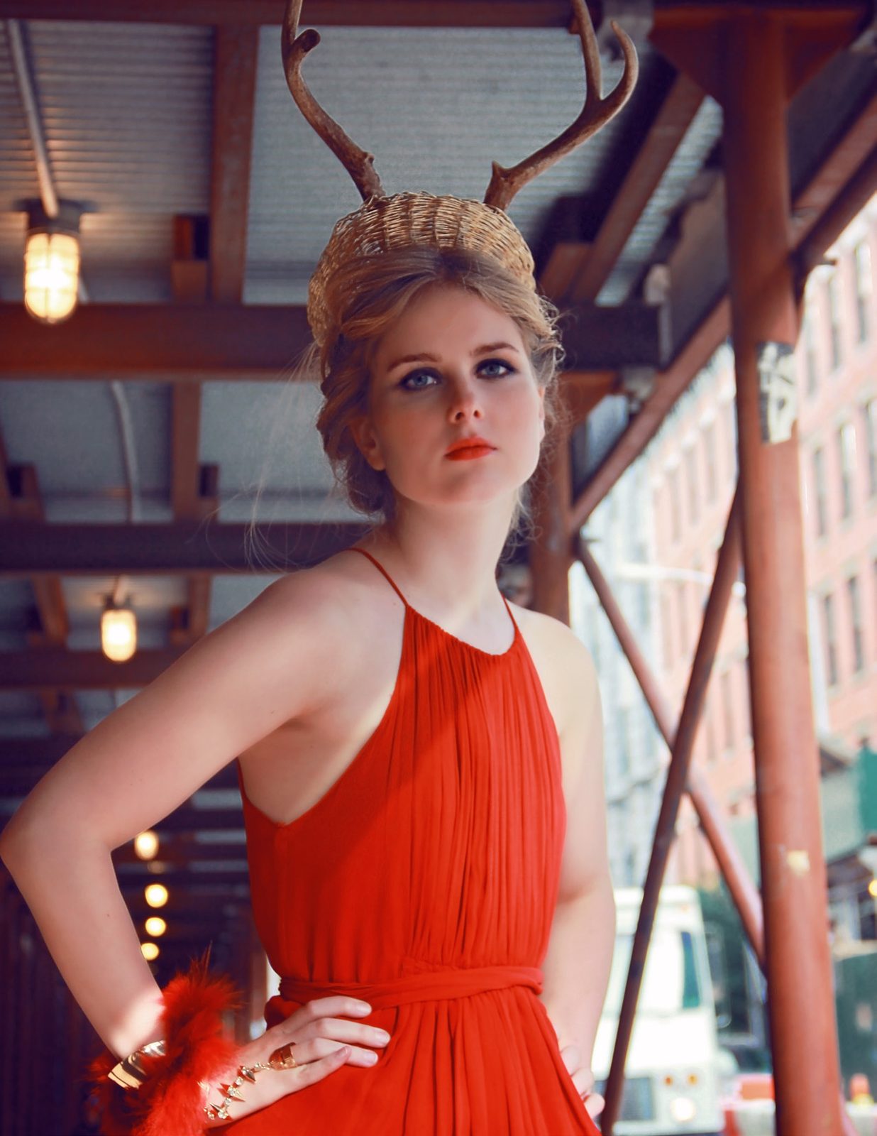 Model in a red halter gown and antler crown stands beneath steel scaffolding, hand on hip