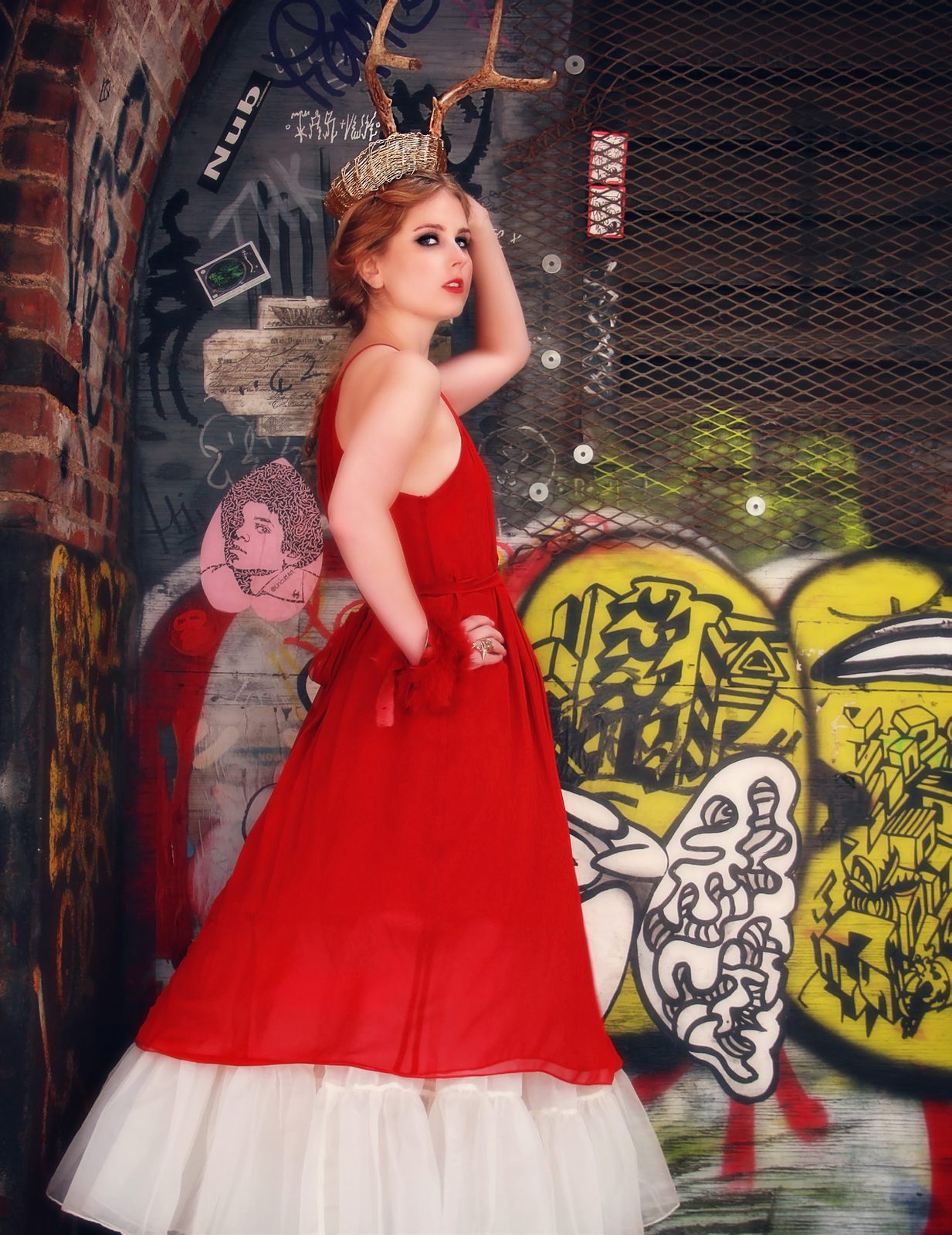 Model in a red gown with a white tulle underskirt poses against a graffiti mural, wearing an antler crown