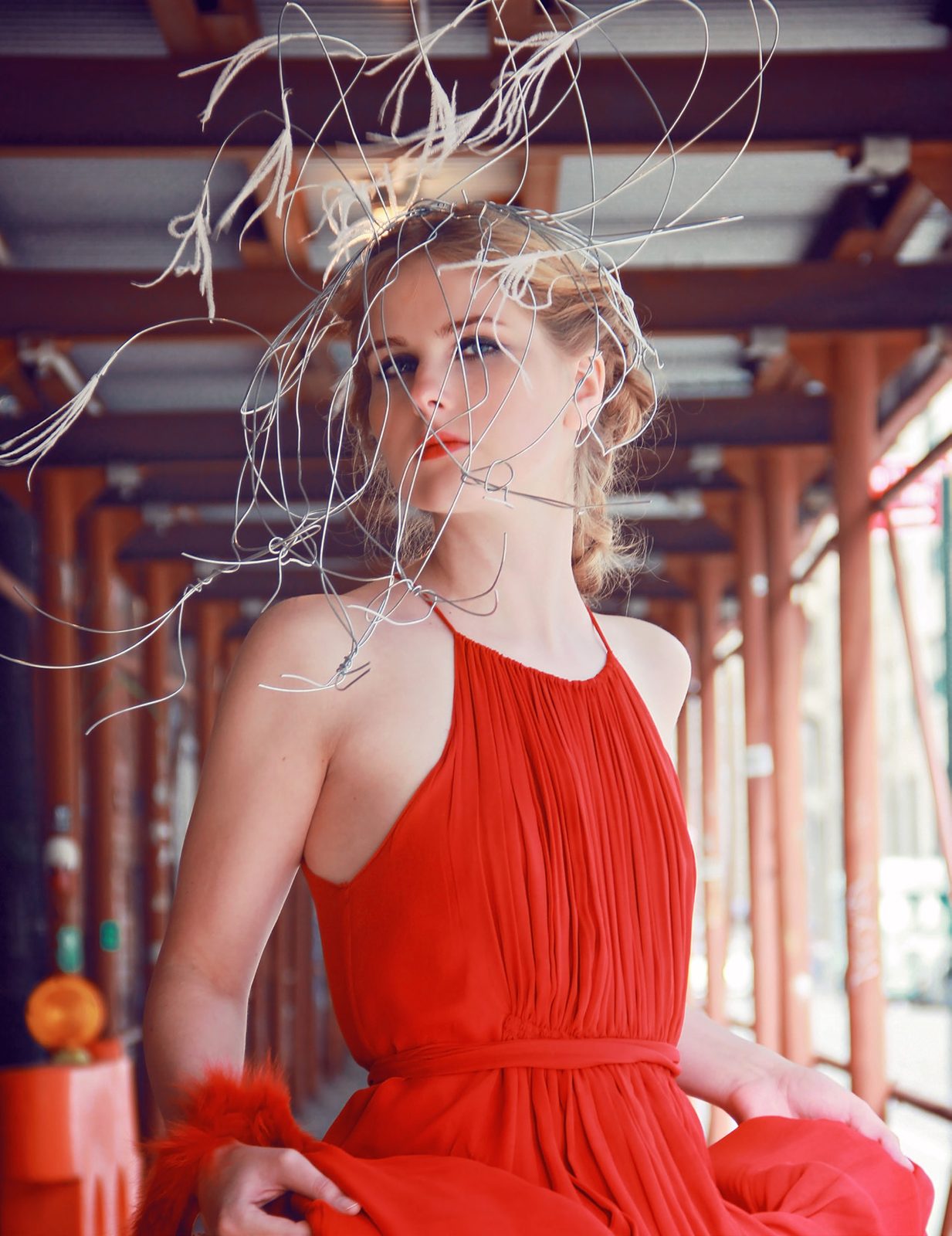 Model in a red halter gown with a wild wire headpiece stands under orange scaffolding