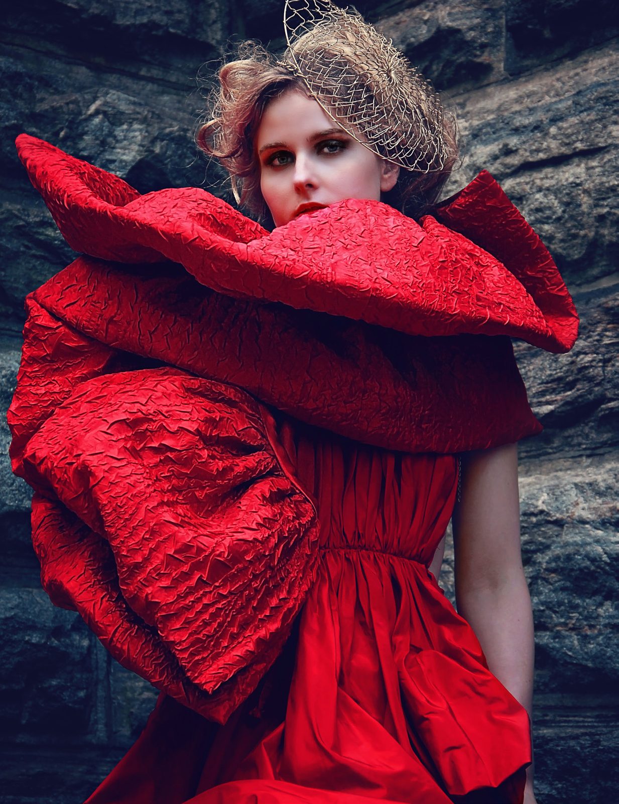 Model wrapped in an enormous sculptural red coat with a caged headpiece against a stone wall