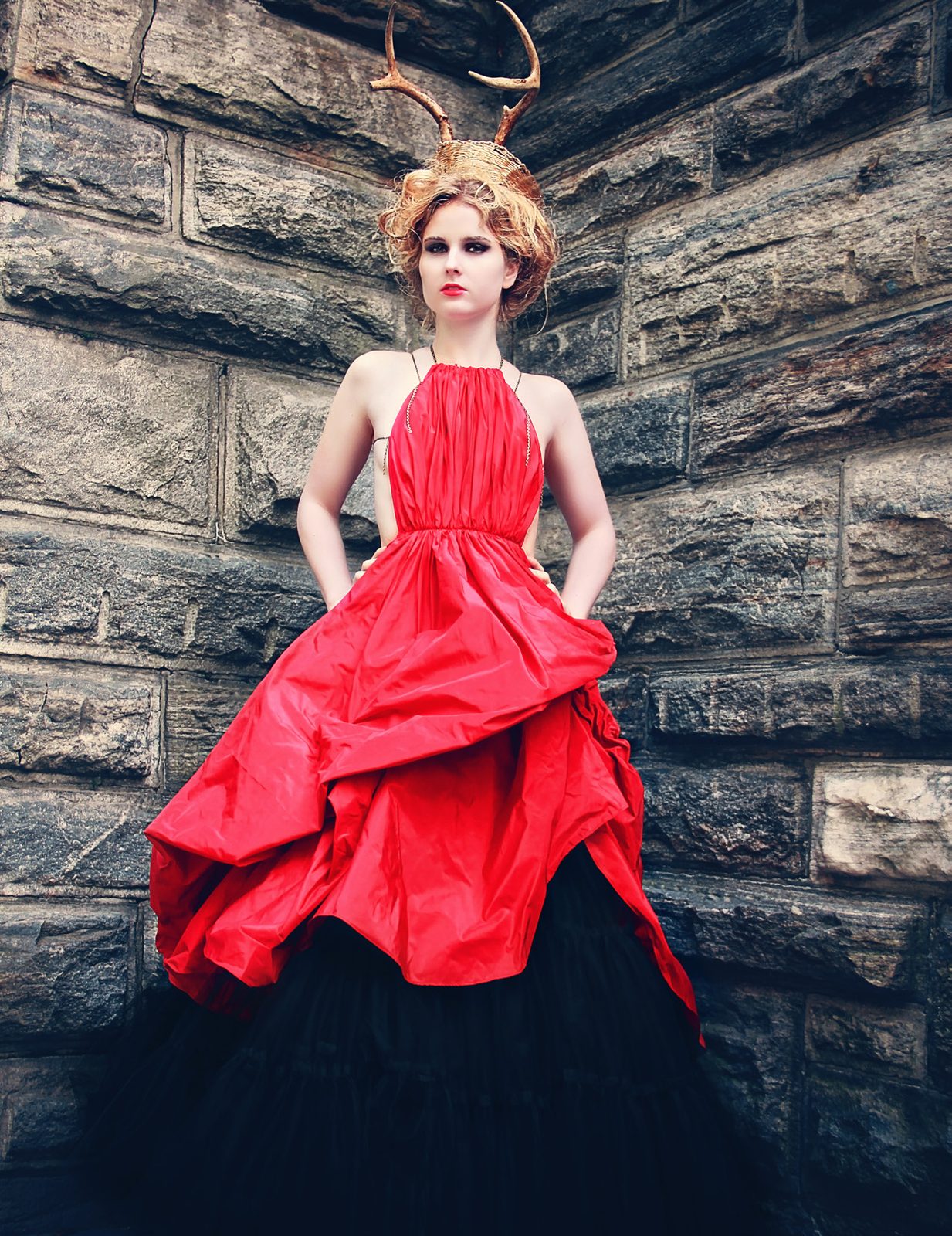 Model in a red gown over a black tulle skirt, antler crown on her head, stands against a stone wall