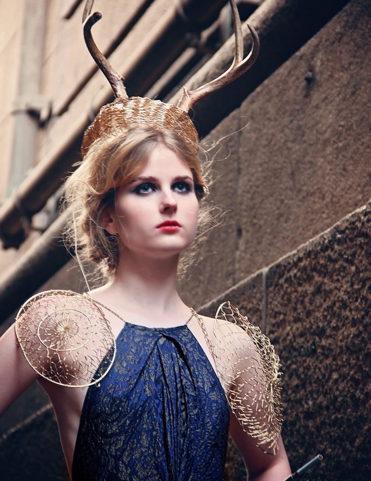 Model in a navy beaded gown with gold wire shoulder pieces and tall antlers in a narrow alley