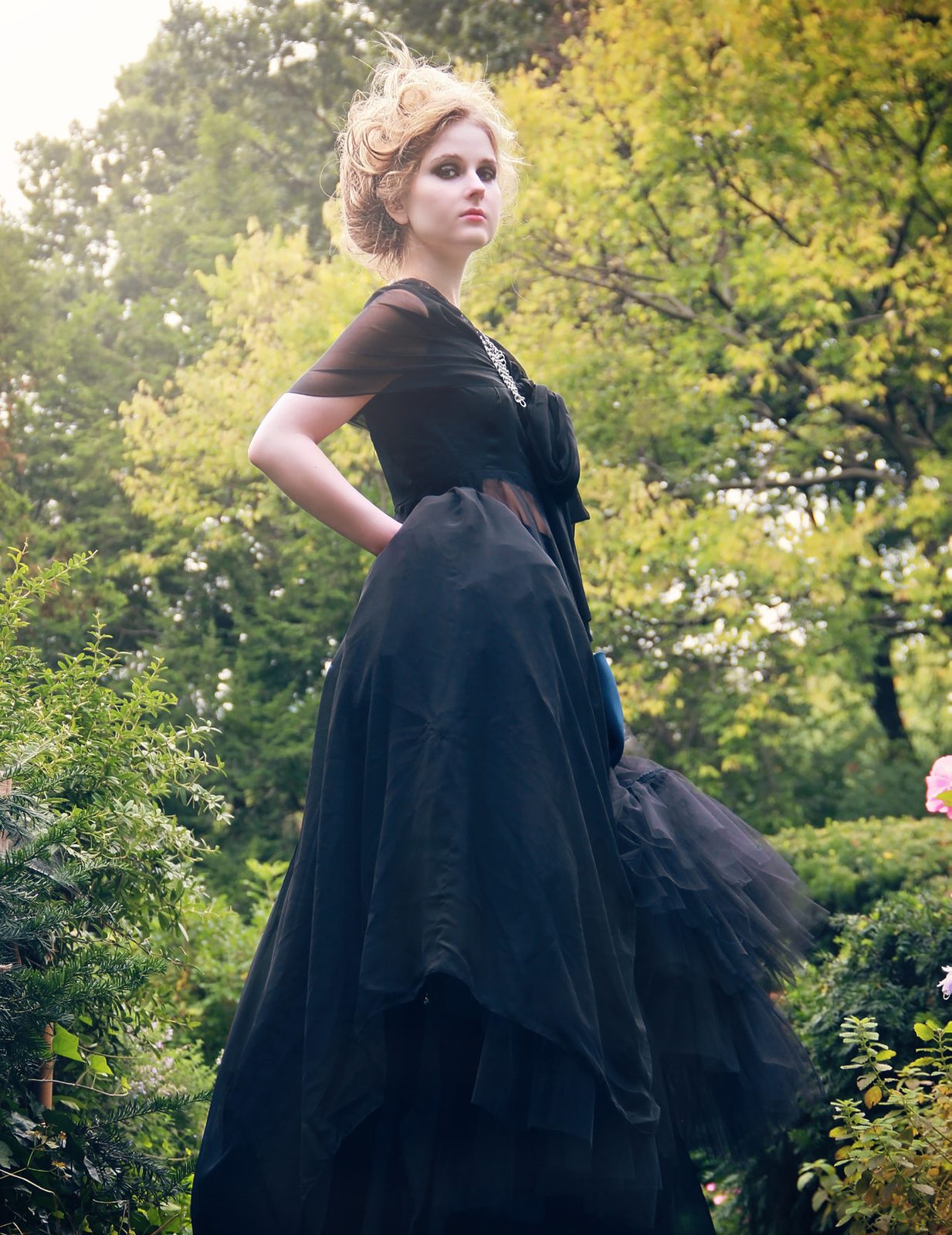Model in a long black gown with a tulle skirt stands in a leafy park