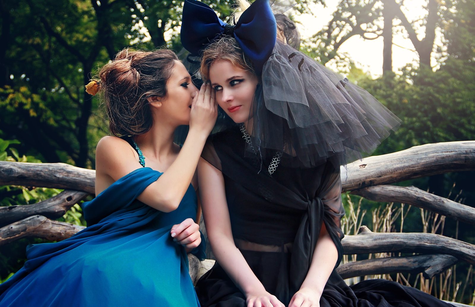 Two models on a rustic wooden bench, one in teal silk whispering to the other in black tulle and a giant navy bow headpiece