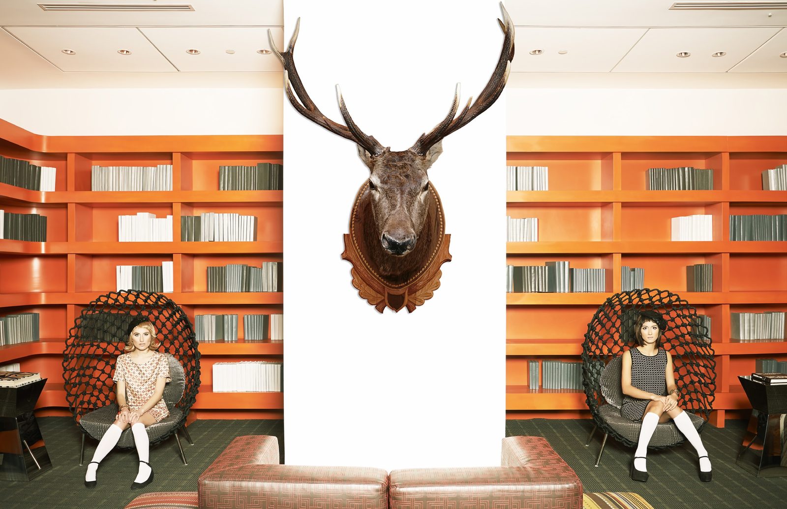 Two models in mod dresses and white knee socks sit in woven pod chairs on either side of a mounted stag head, framed by orange bookshelves
