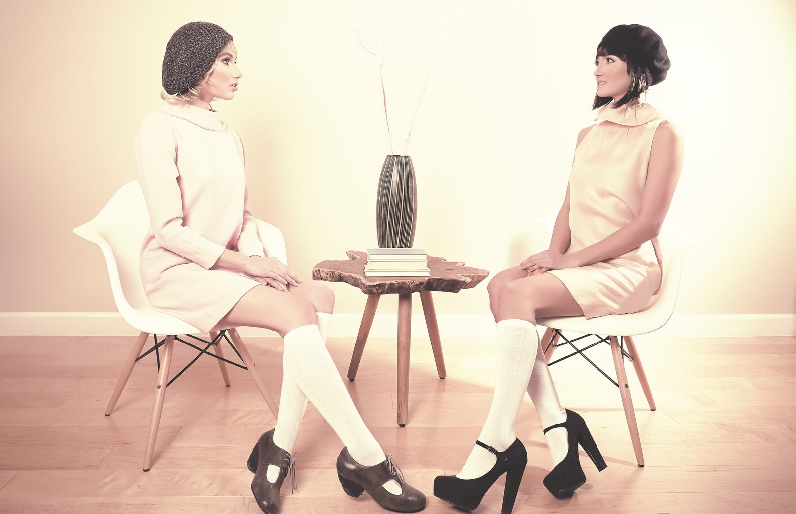 Two models in pastel mod shift dresses, knee socks and berets sit facing each other across a small wooden table