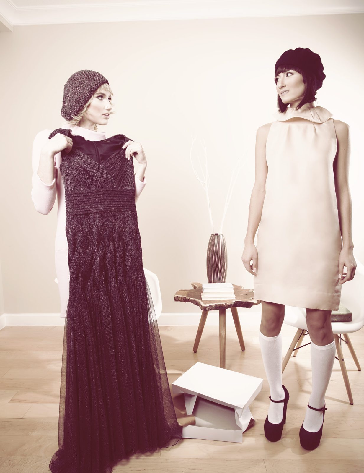 Models in berets hold up a long wine-colored gown and a pale shift dress in a minimal white room
