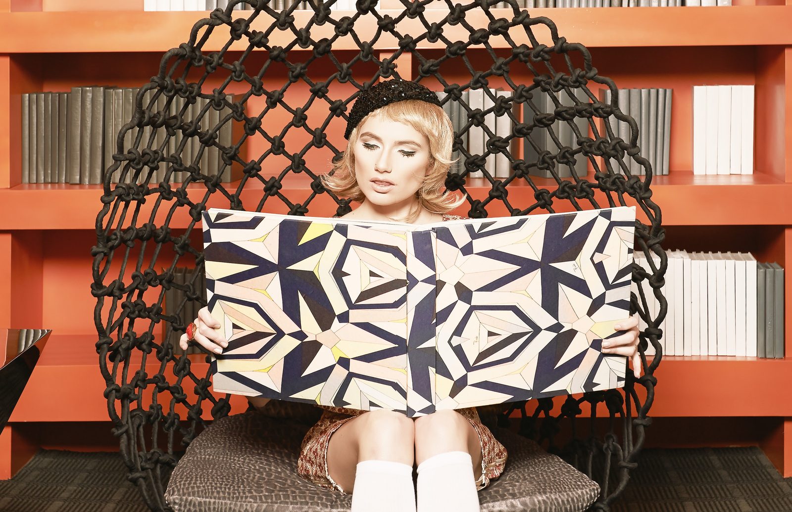 Model in a pod chair holds open a geometric print book in front of orange bookshelves