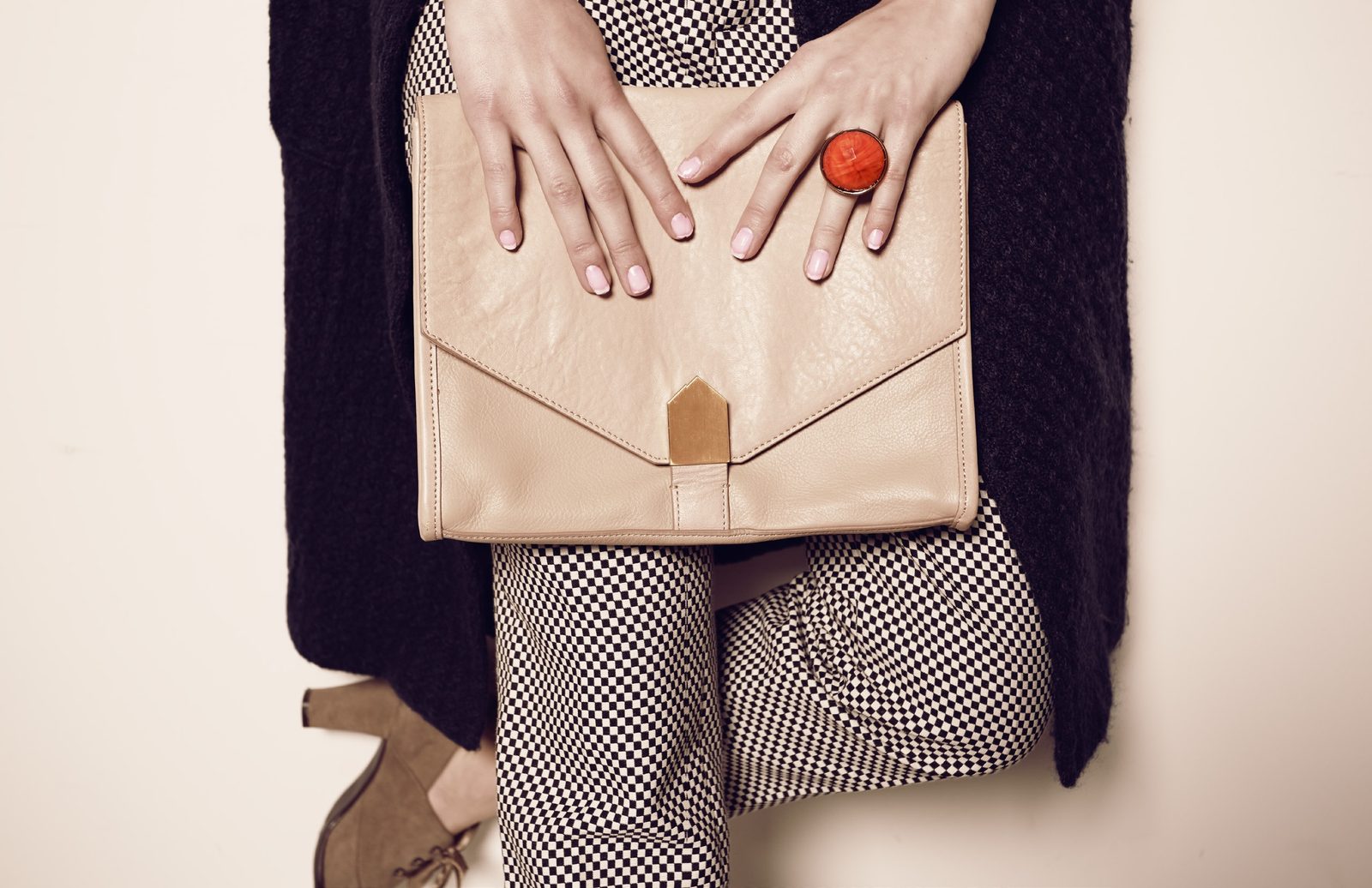 Close crop of hands with a red cocktail ring resting on a nude leather envelope clutch over houndstooth pants