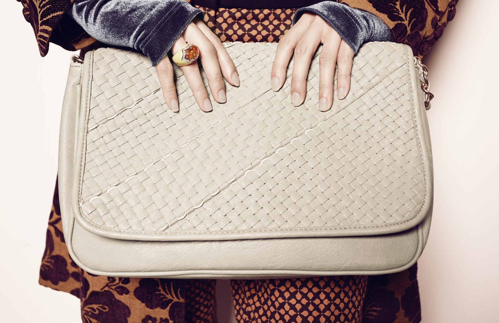 Close crop of hands resting on a woven cream leather crossbody bag over a velvet floral jacket