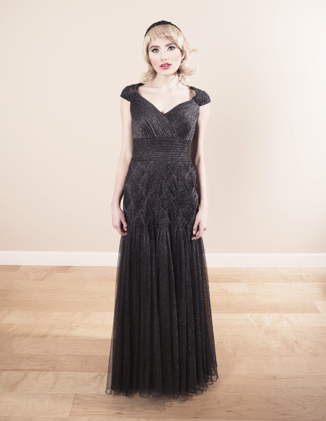 Model in a long black cap-sleeve tulle gown and headband stands against a blush wall