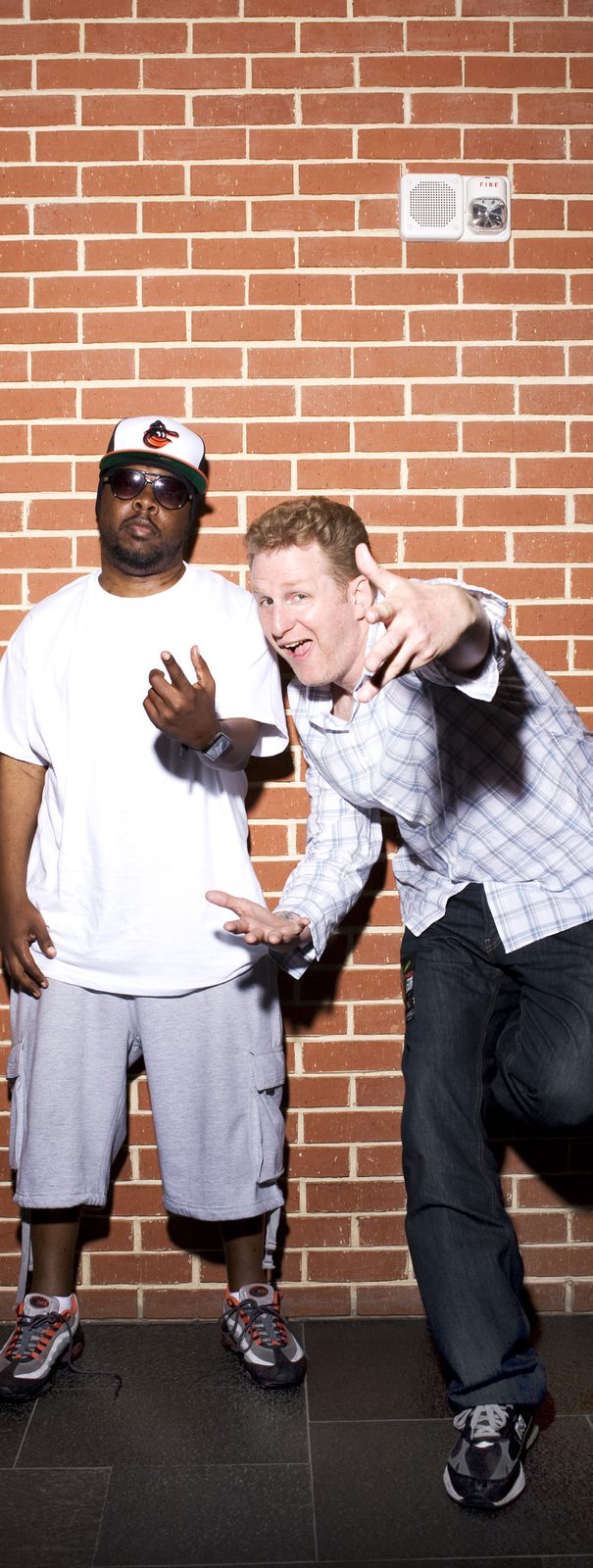 Phife Dawg in a white tee and baseball cap standing beside Michael Rapaport in a plaid shirt against a brick wall