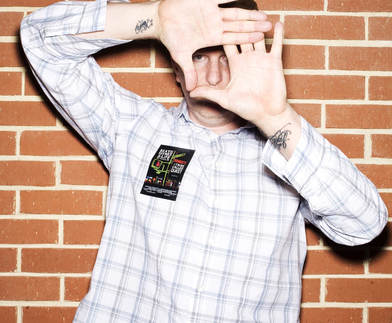 Michael Rapaport in a plaid shirt holding his hands up in front of his face against a brick wall