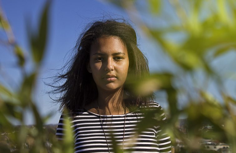 Young woman with windswept dark hair in a striped top, standing among tall green grass under a blue sky