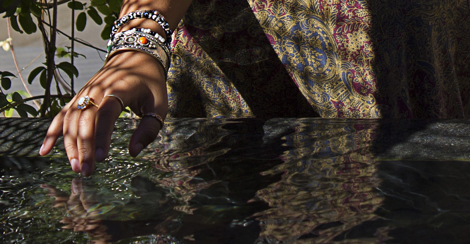 A hand stacked with beaded bracelets and rings trails its fingertips across dark water