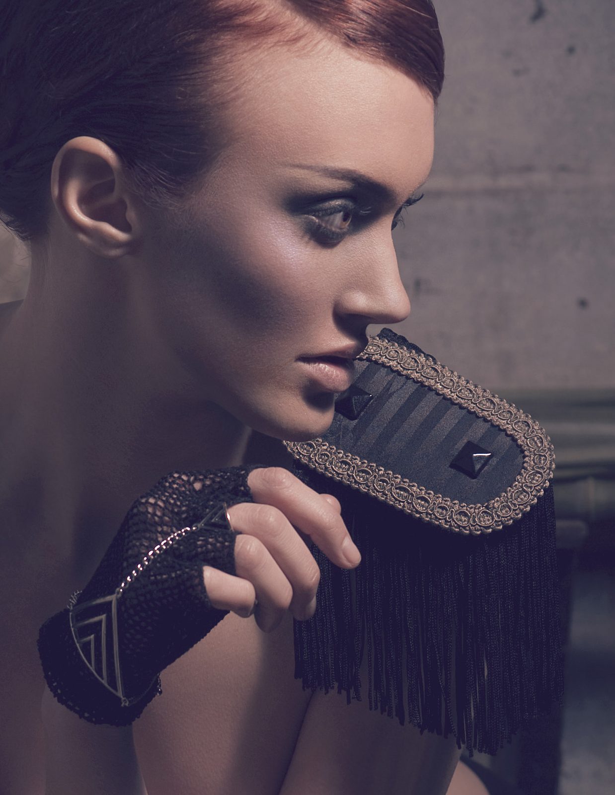 Model with swept copper hair and smoky eyes in a fringed black epaulet and fingerless leather glove, looking over her shoulder