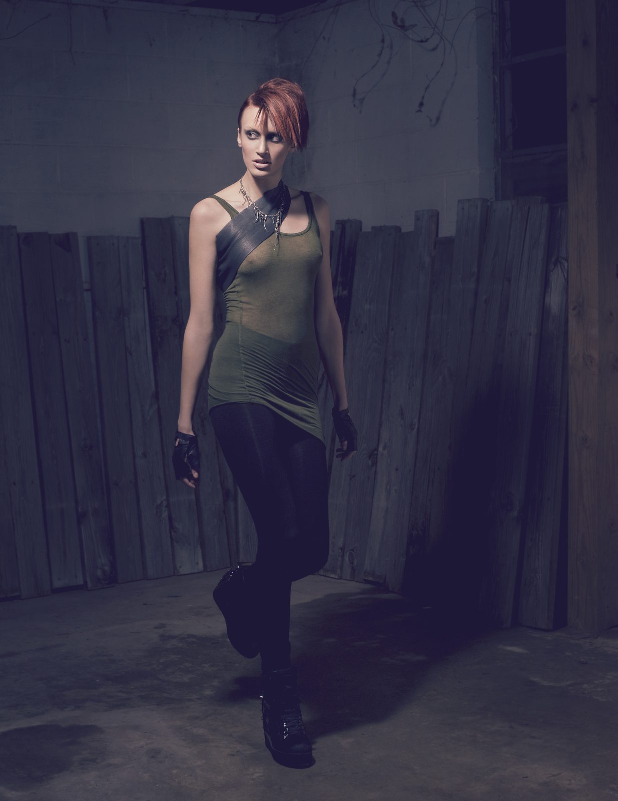 Model walking through a dim concrete room in an olive tank, black leggings, boots and a leather shoulder strap