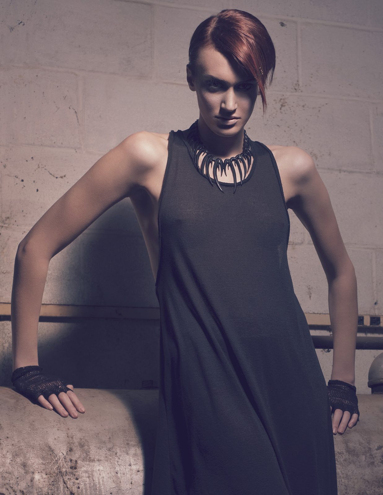 Model in a long charcoal sleeveless dress and a spiked statement collar necklace, leaning on a concrete ledge