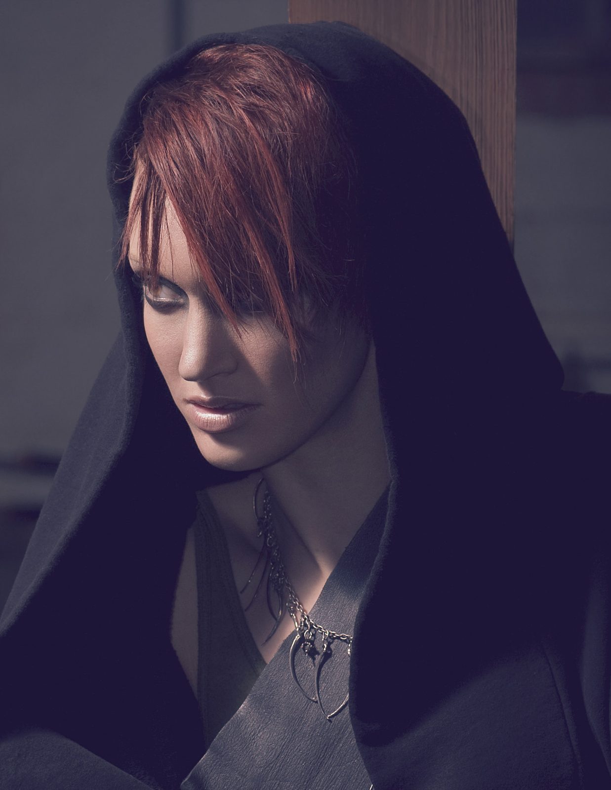 Model in a deep black hood looking down, a spiked silver necklace at her chest